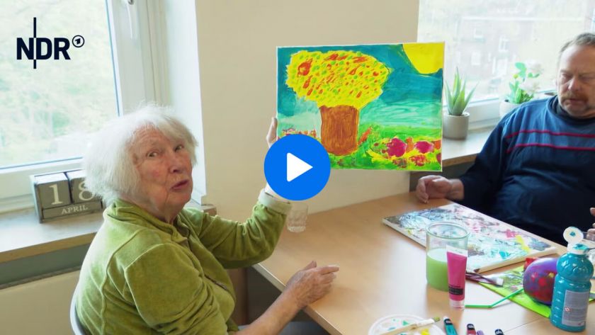 Video Screenshot, NDR über zusammenkultur, eine ältere Frau hebt ein selbstgemaltes Bild hoch, ein Mann sitzt am Tisch daneben und schaut zu