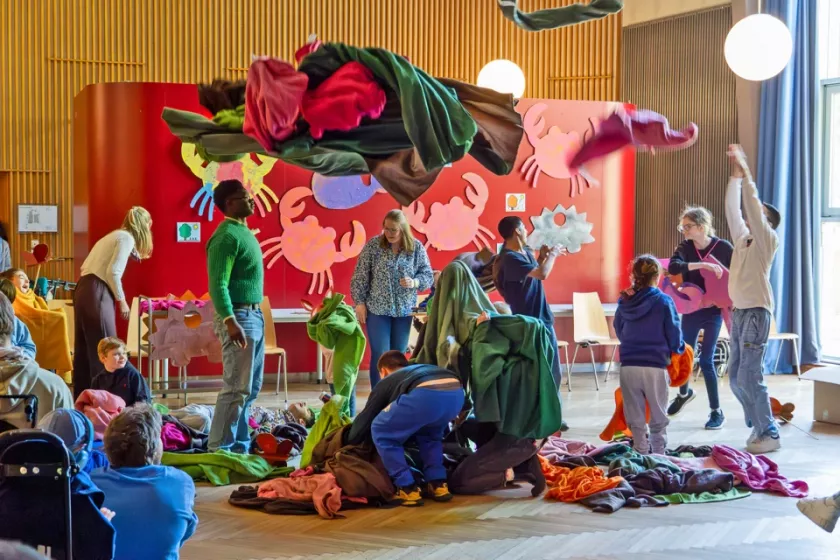 Kinder, Jugendliche und Erwachsene mit und ohne Behinderung bewegen gemeinsam farbige Decken während einer Szene des inklusiven Theaterprojekts Decken.Farben.Jahreszeiten in einem großen Veranstaltungsraum.