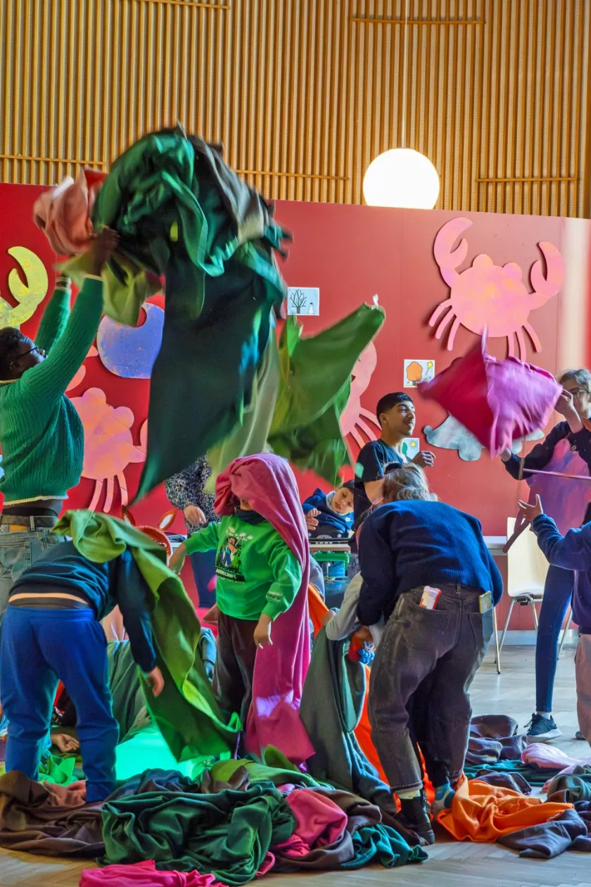 Kinder, Jugendliche und Erwachsene mit und ohne Behinderung bewegen gemeinsam farbige Decken während einer Szene des inklusiven Theaterprojekts Decken.Farben.Jahreszeiten in einem großen Veranstaltungsraum.
